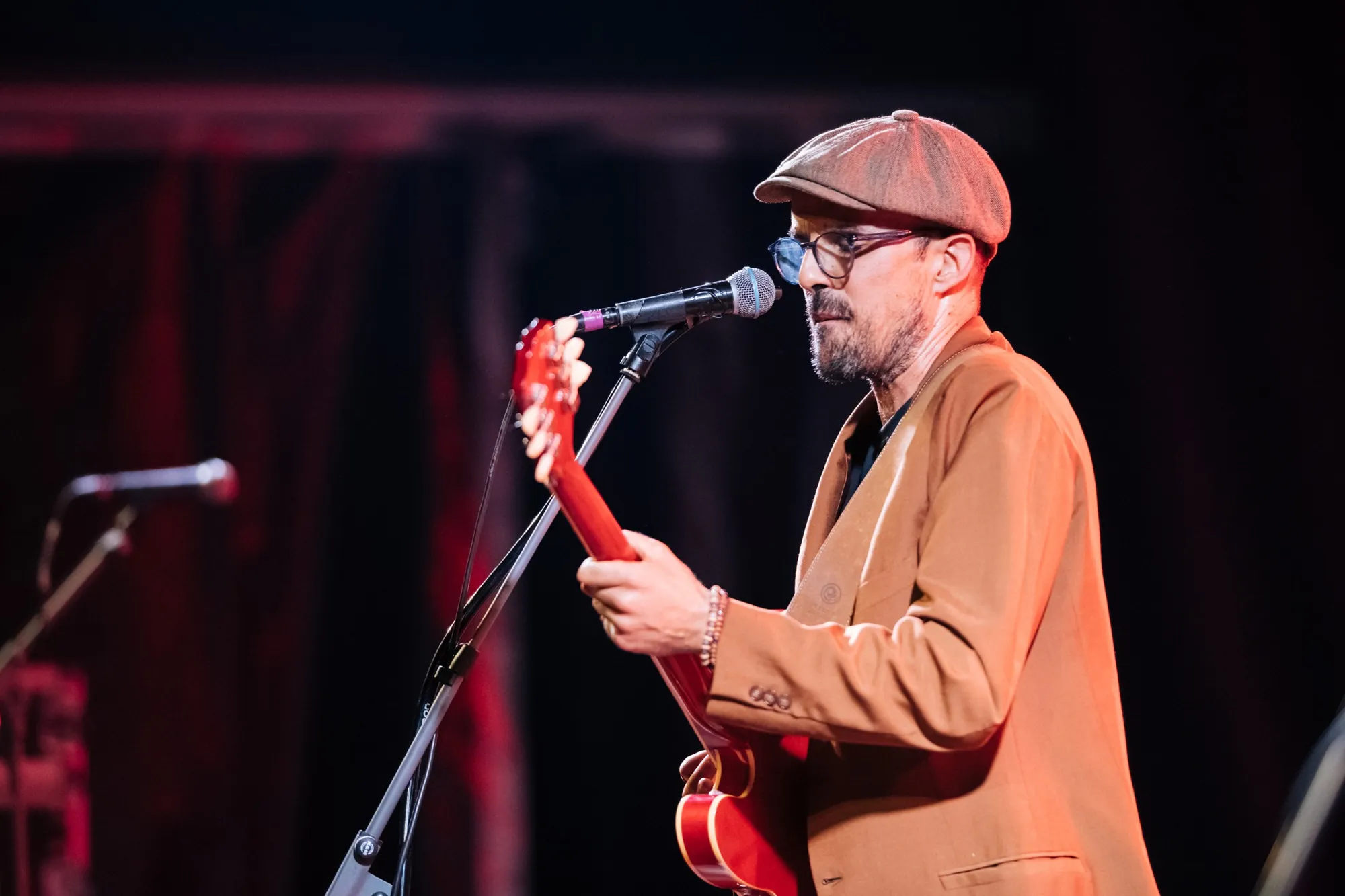 Músico de boina e casaco laranja canta e toca guitarra no palco do MeaJazz